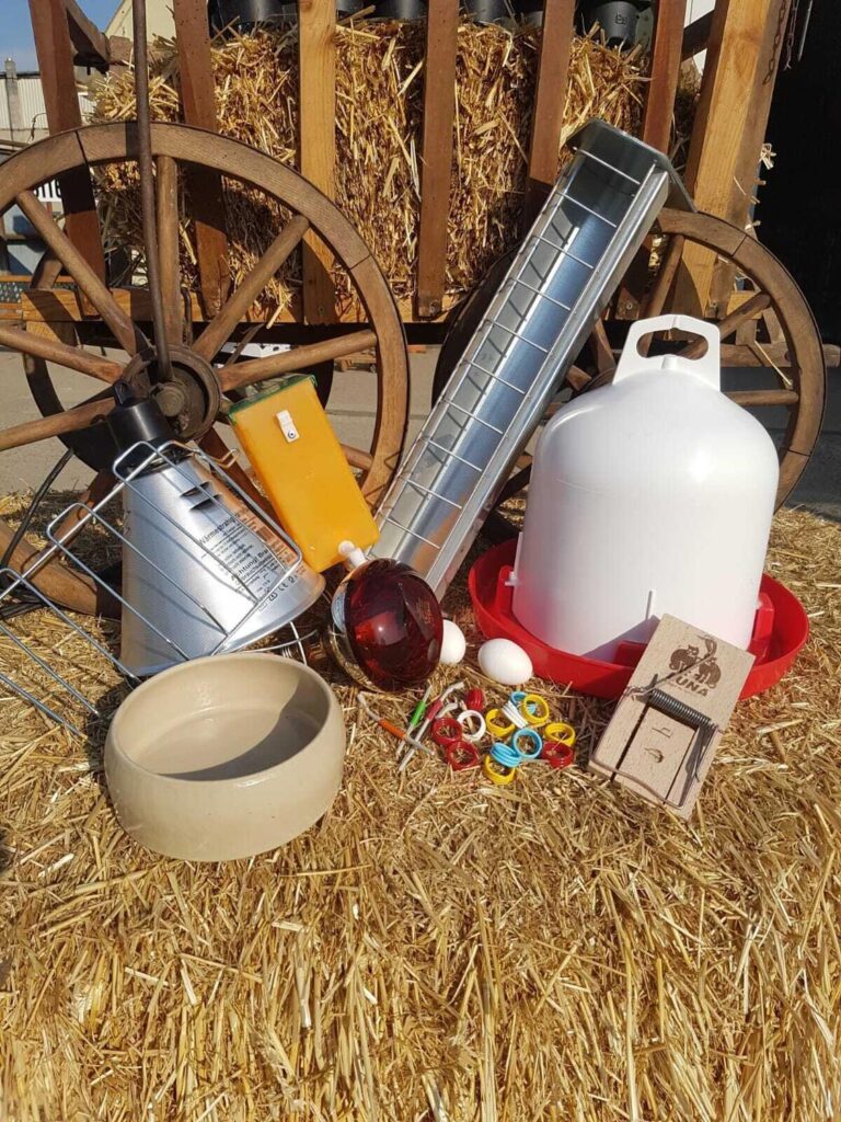 Ein Haufen bunter und silberner Hühnerzubehörteile liegt auf Stroh vor einem Holzwagen